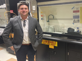 A professionally dressed man smiling at the camera, standing next to a scientific lab space.