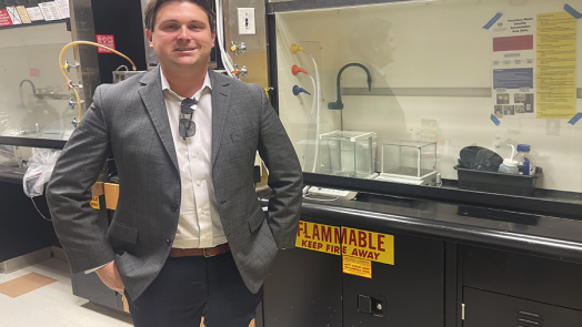 A professionally dressed man smiling at the camera, standing next to a scientific lab space.