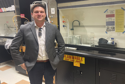 A professionally dressed man smiling at the camera, standing next to a scientific lab space.