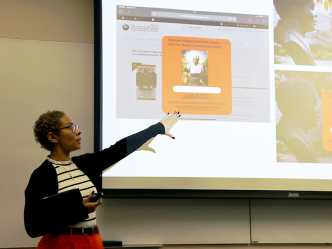 A female college student stands at the front of a classroom and presents to her class. She is pointing to projection screen with information about a non-profit organization.