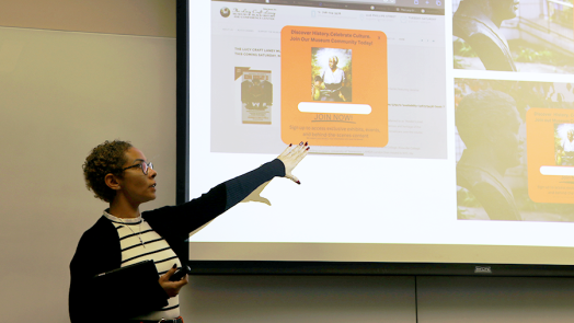 A female college student stands at the front of a classroom and presents to her class. She is pointing to projection screen with information about a non-profit organization.