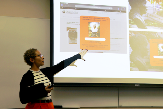 A female college student stands at the front of a classroom and presents to her class. She is pointing to projection screen with information about a non-profit organization.