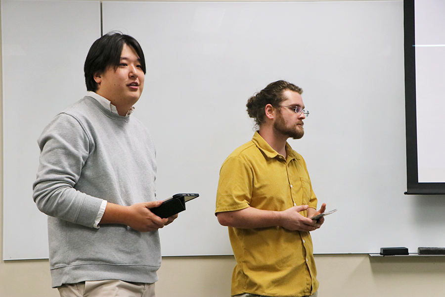 Two male college students present at the front of a class during a project. One is holding an electronic tablet.