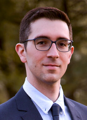 A man in glasses wearing a suit and tie stands outside.