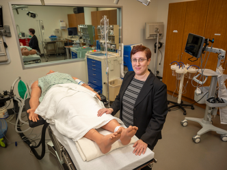 A professionally dressed person stands beside a medical manikin in a clinical operating room and smiles at the camera.