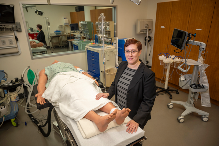 A professionally dressed person stands beside a medical manikin in a clinical operating room and smiles at the camera.