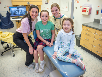 Two female dentists and two female children smile at the camera. They are in a dental exam room, with an X-ray on the screen behind them.