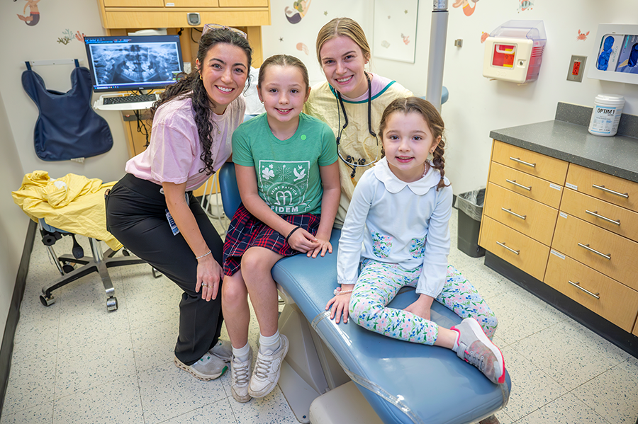 Two female dentists and two female children smile at the camera. They are in a dental exam room, with an X-ray on the screen behind them.