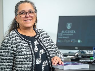 a woman stands at a desk with a computer with the Augusta University logo on the screen behind her