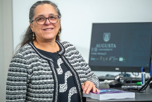 a woman stands at a desk with a computer with the Augusta University logo on the screen behind her
