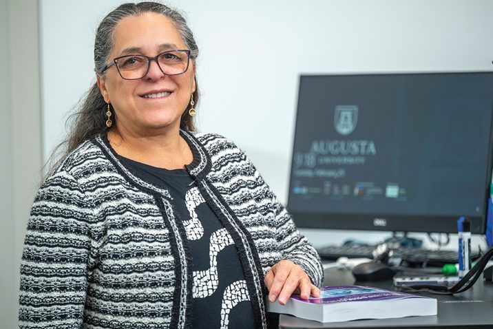 a woman stands at a desk with a computer with the Augusta University logo on the screen behind her