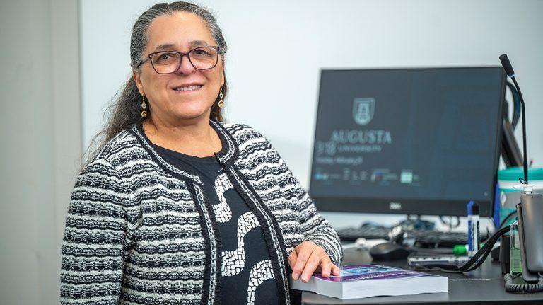 a woman stands at a desk with a computer with the Augusta University logo on the screen behind her