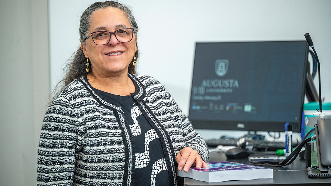 a woman stands at a desk with a computer with the Augusta University logo on the screen behind her