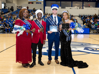 Two college men and two college women, all wearing crowns and sashes, smiling in a basketball gymnasium.