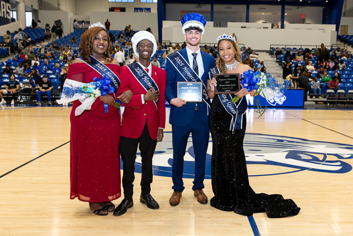 Two college men and two college women, all wearing crowns and sashes, smiling in a basketball gymnasium.