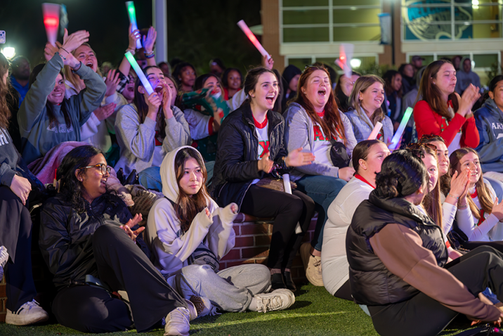 College students sit around an outdoor amphitheater and cheer during a Homecoming event.