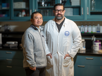 Two men stand in a scientific lab. One man is wearing a lab coat.