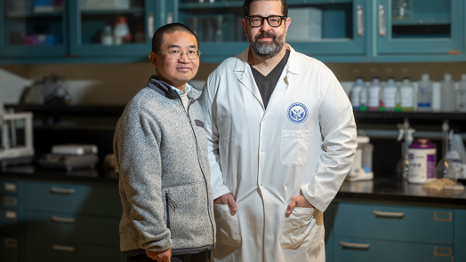 Two men stand in a scientific lab. One man is wearing a lab coat.