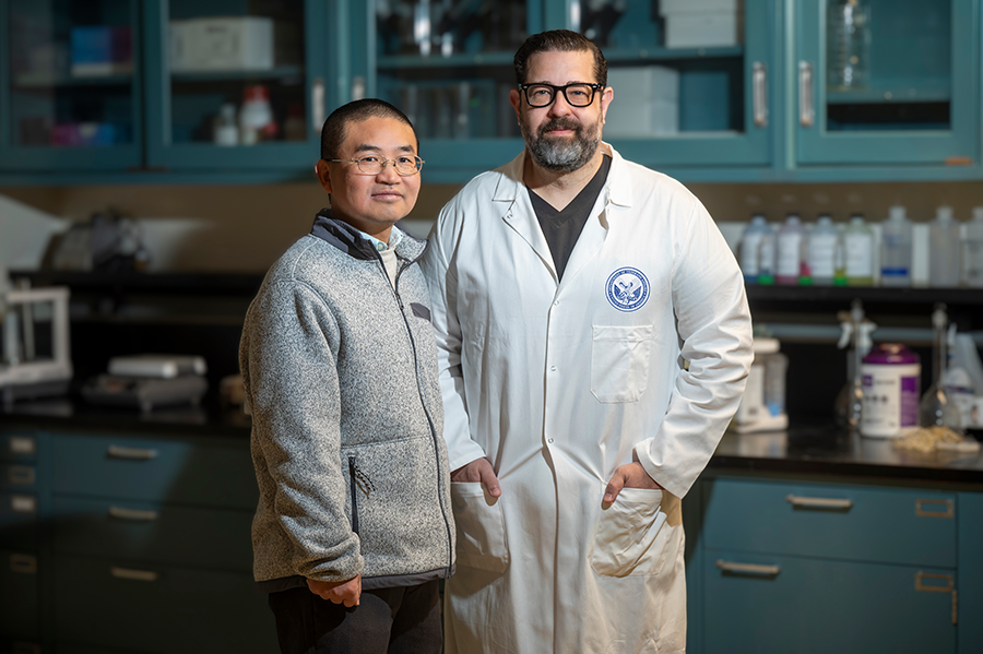 Two men stand in a scientific lab. One man is wearing a lab coat.