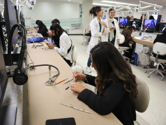 A young woman sits at a desk and practices using a dental pick on a piece of plaster. There are dental students and other people in the background doing the same thing.