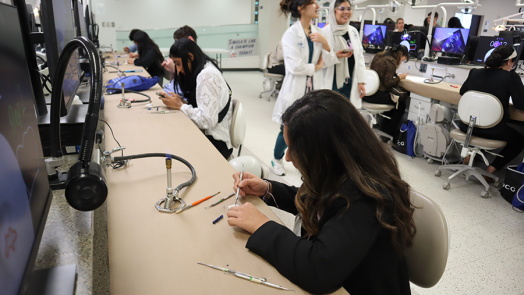 A young woman sits at a desk and practices using a dental pick on a piece of plaster. There are dental students and other people in the background doing the same thing.