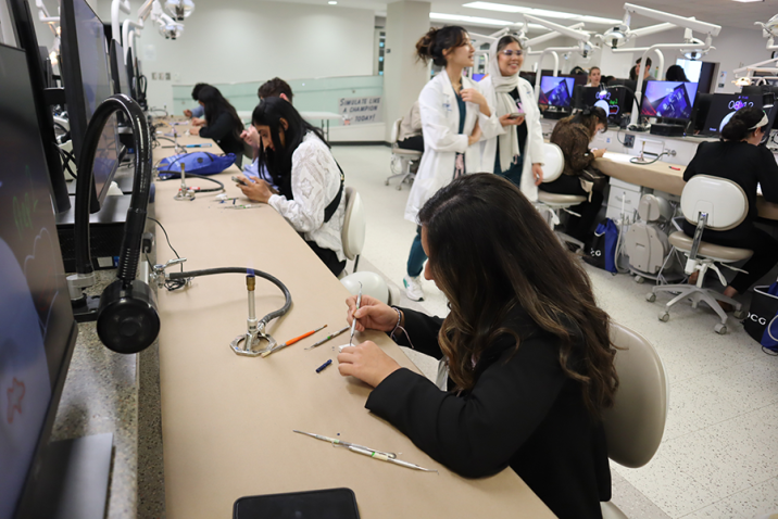 A young woman sits at a desk and practices using a dental pick on a piece of plaster. There are dental students and other people in the background doing the same thing.