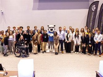 A large group of people, men and women, pose in front of banners with school mascot wearing big cat costume head.