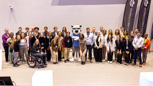 A large group of people, men and women, pose in front of banners with school mascot wearing big cat costume head.