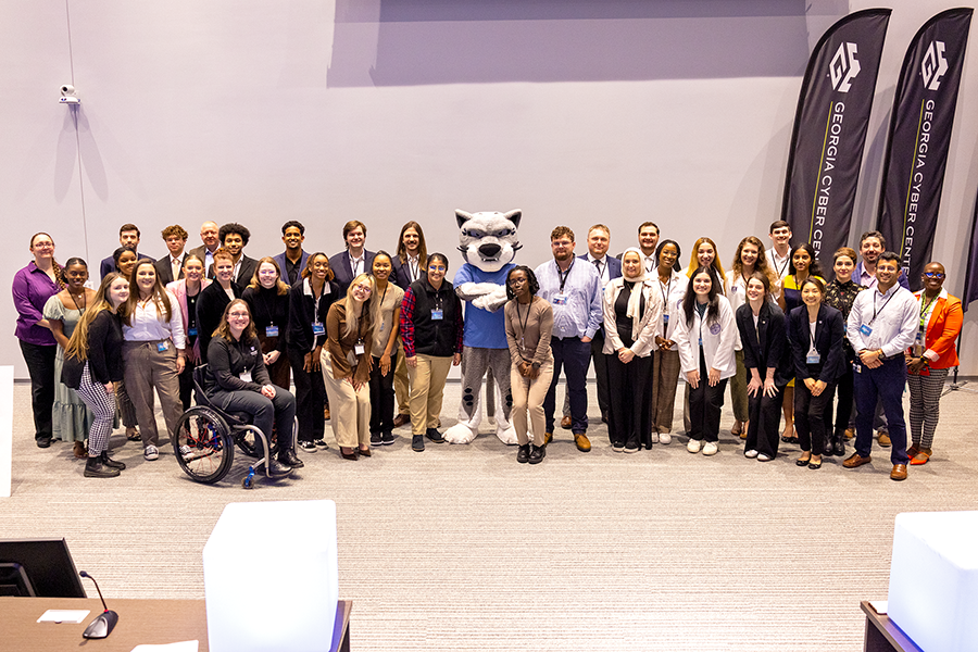 A large group of people, men and women, pose in front of banners with school mascot wearing big cat costume head.