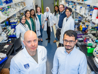 A group of ten scientists in lab coats smile at the camera. There are shelves of materials for experiments around them. Two scientists stand in front of the others.