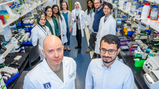 A group of ten scientists in lab coats smile at the camera. There are shelves of materials for experiments around them. Two scientists stand in front of the others.