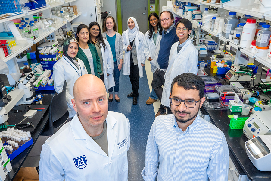 A group of ten scientists in lab coats smile at the camera. There are shelves of materials for experiments around them. Two scientists stand in front of the others.