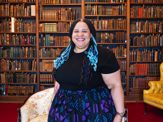 A woman stands in a large library with books all around. There is a love seat behind her and an arm chair in one corner of the room.