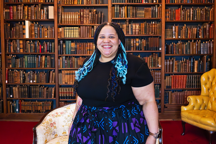 A woman stands in a large library with books all around. There is a love seat behind her and an arm chair in one corner of the room.