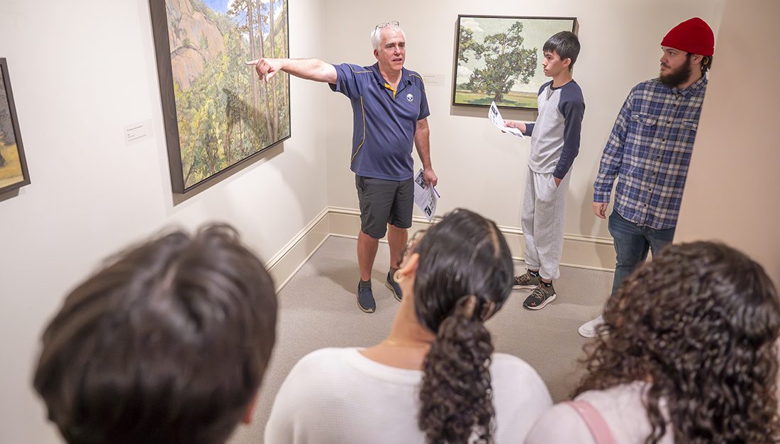 A man stands near a piece of art and points to it as he talks to a group of students about it