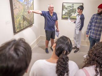 A man stands near a piece of art and points to it as he talks to a group of students about it