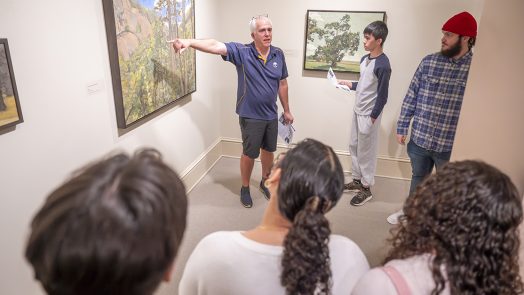 A man stands near a piece of art and points to it as he talks to a group of students about it