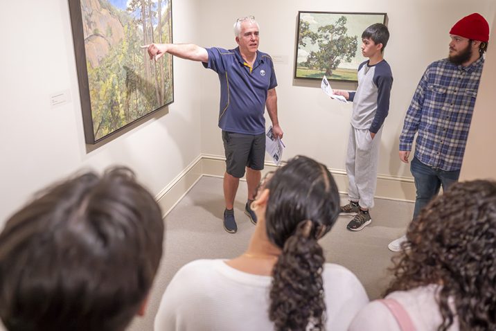 A man stands near a piece of art and points to it as he talks to a group of students about it