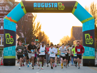 Runners start a race. There is a large inflatable arch marking the start line. A logo for the Augusta Sports Council is featured at the top.