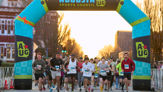 Runners start a race. There is a large inflatable arch marking the start line. A logo for the Augusta Sports Council is featured at the top.