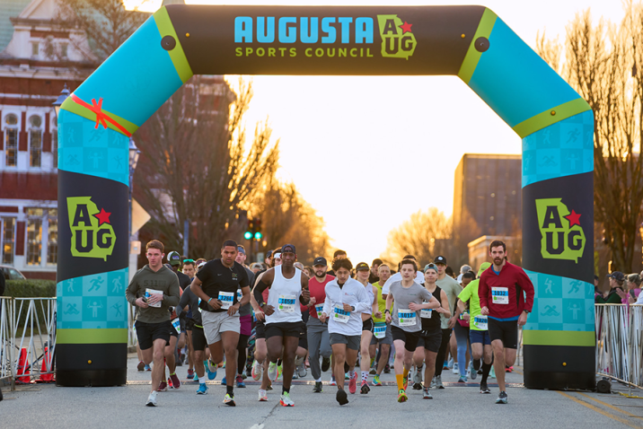 Runners start a race. There is a large inflatable arch marking the start line. A logo for the Augusta Sports Council is featured at the top.