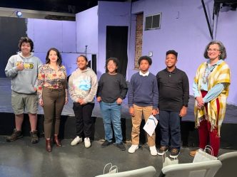 A group of five middle school students stand on a stage with two female college professors.