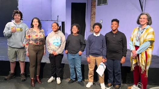 A group of five middle school students stand on a stage with two female college professors.
