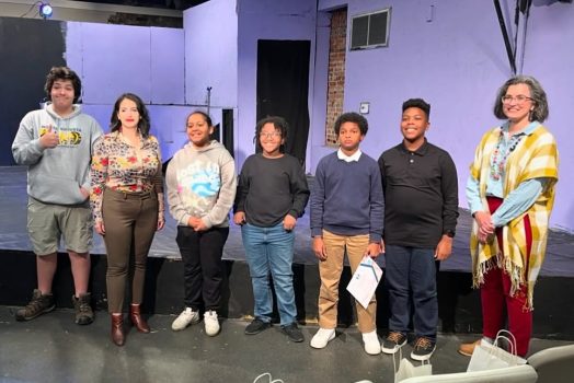 A group of five middle school students stand on a stage with two female college professors.