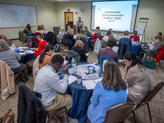 About 25 people sit at circular tables in a conference room. A man stands at the front by a large projector screen with the words "Case Scenario: Giving Constructive Feedback" on it.