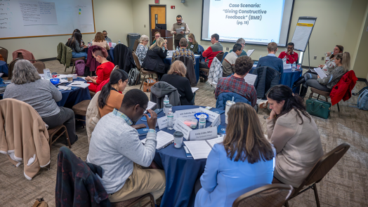 About 25 people sit at circular tables in a conference room. A man stands at the front by a large projector screen with the words "Case Scenario: Giving Constructive Feedback" on it.