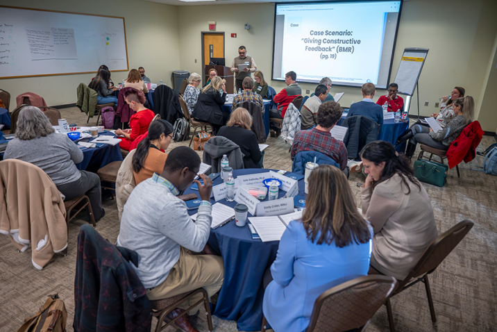 About 25 people sit at circular tables in a conference room. A man stands at the front by a large projector screen with the words "Case Scenario: Giving Constructive Feedback" on it.