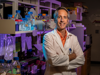 A scientist in a lab coat stands in front of a table in a research lab and smiles at the camera.