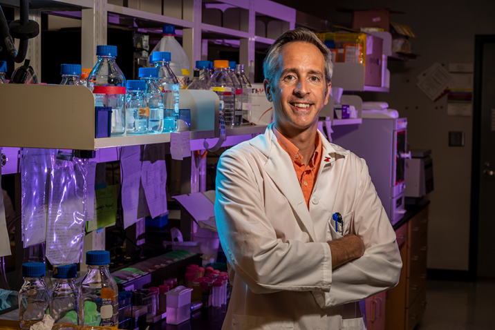 A scientist in a lab coat stands in front of a table in a research lab and smiles at the camera.