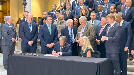 A large group of people gather around a man and a woman as the man signs a piece of paper. They are in a large atrium of a state capitol building.